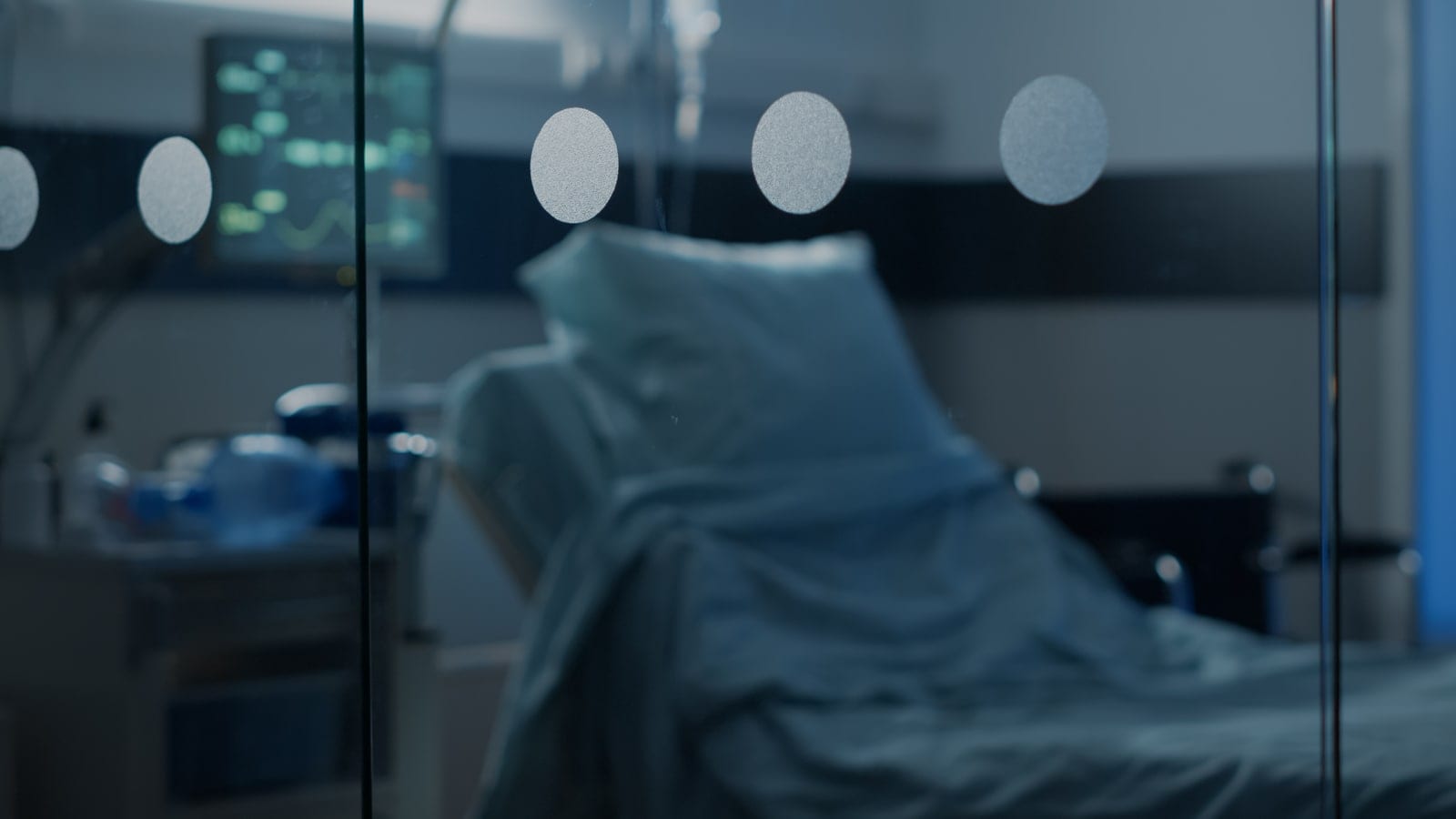 Empty hospital ward bed beside a monitor in low light